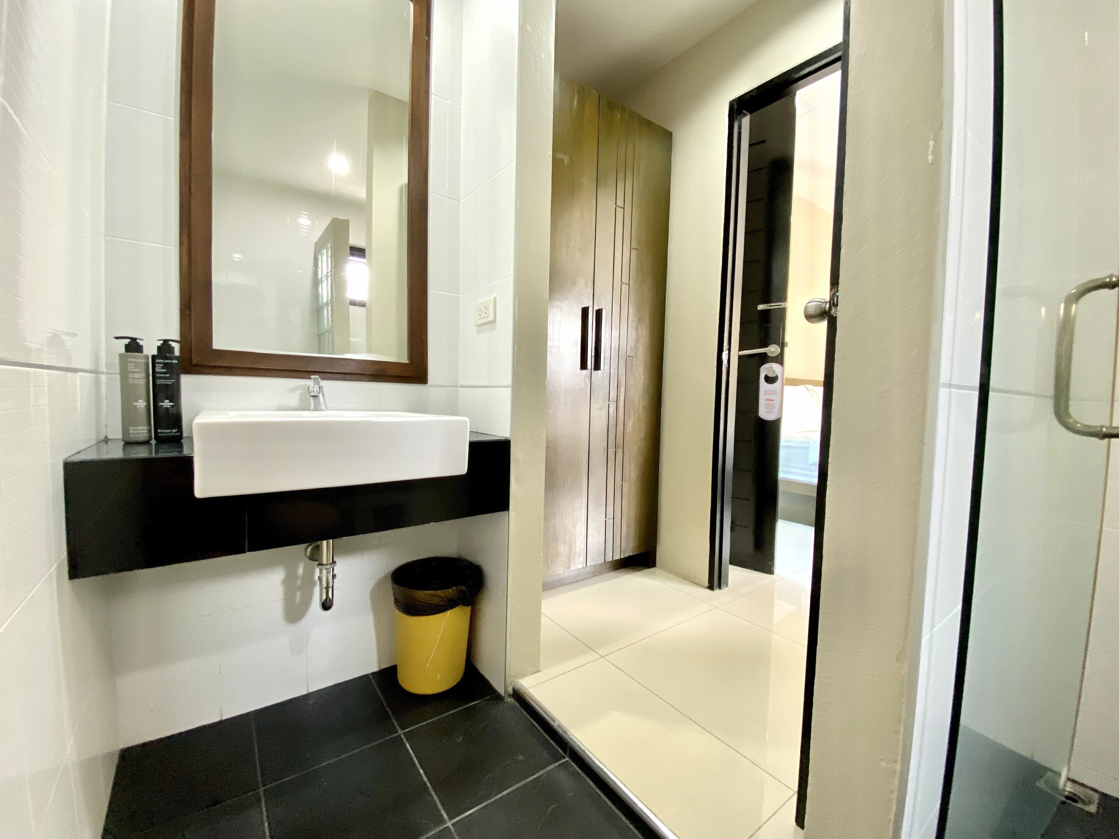 A modern bathroom with a white sink, black countertop, and a mirror above it. There is a trash can and a shower area visible.