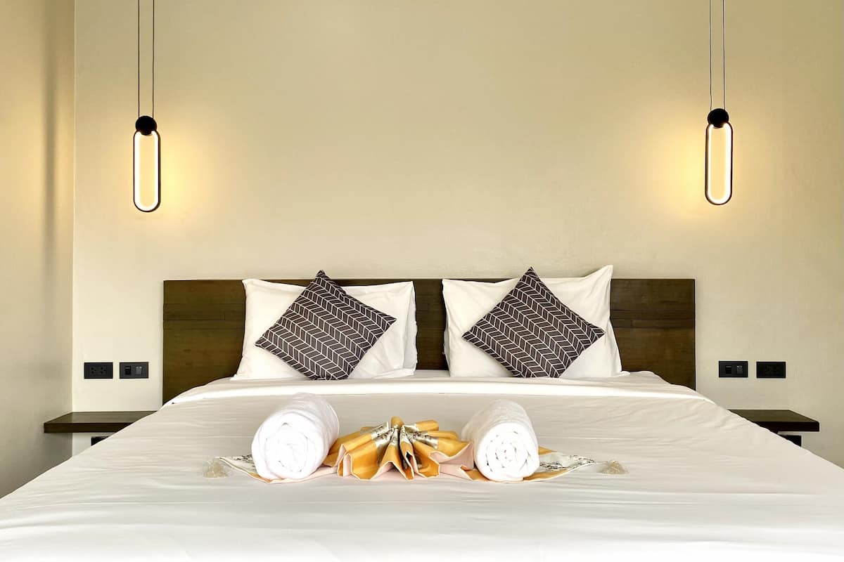 A hotel room with a neatly made bed, two pillows with a geometric pattern, a pair of rolled towels, and two bedside tables with lamps.