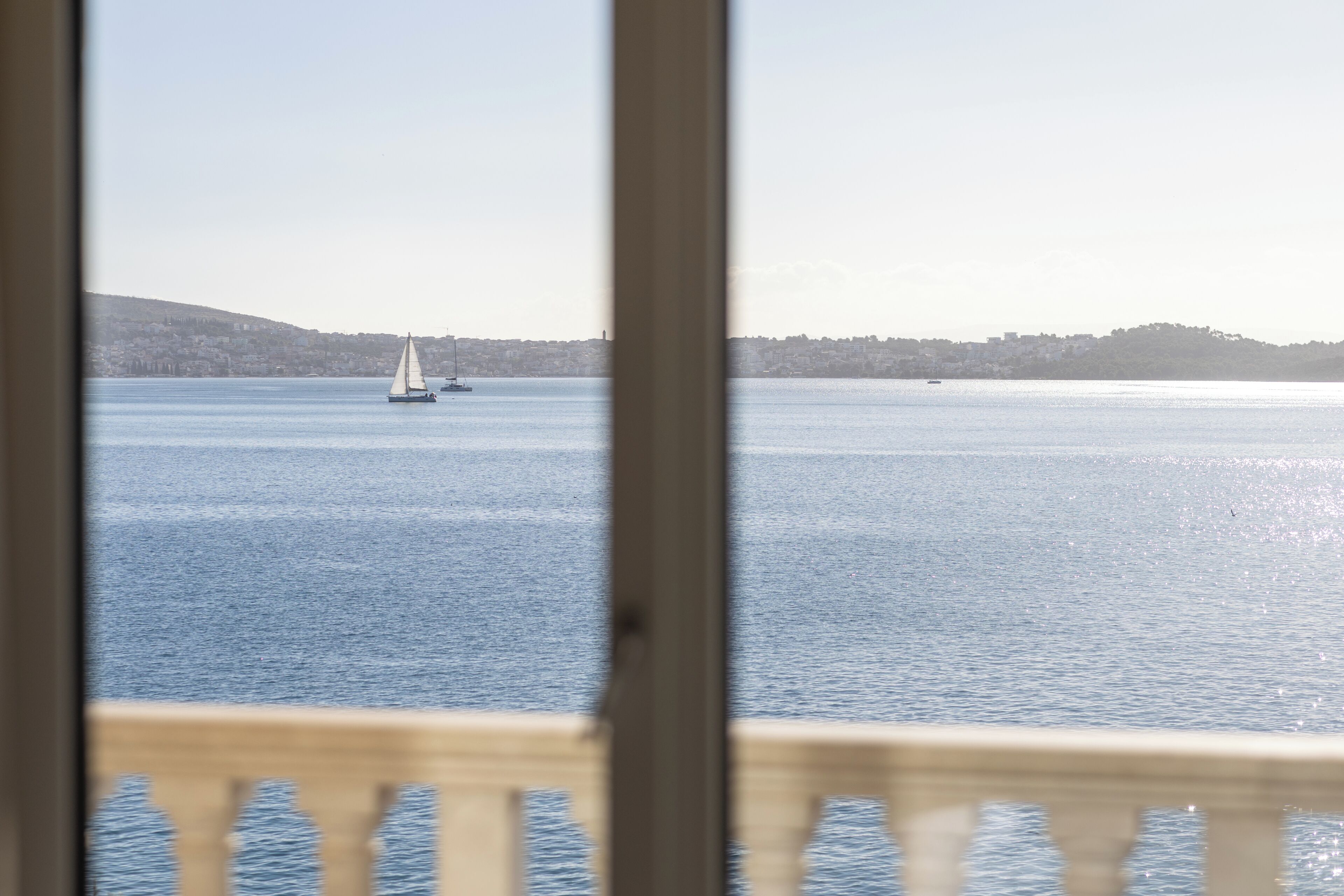 A view of a calm body of water with a sailboat in the distance and a hilly landscape in the background.