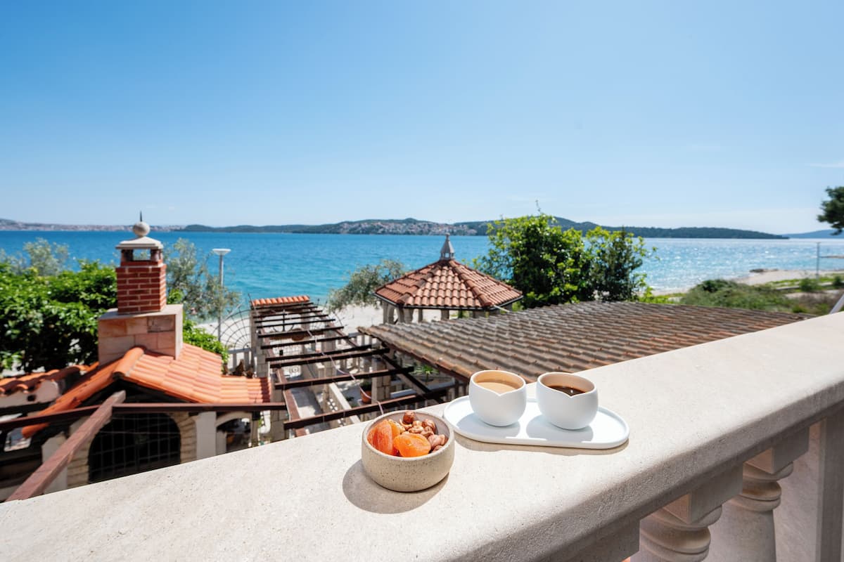 A balcony with a view of the sea, a bowl of fruit, and two cups of coffee on a tray.