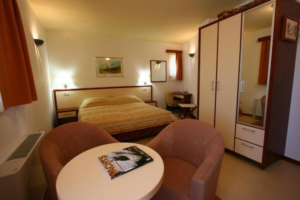 A hotel room with a bed, a desk, a chair, a table with a magazine, a wardrobe, and a mirror.