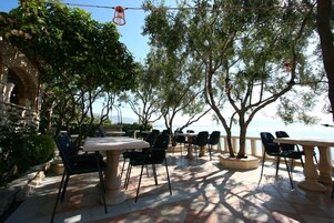 An outdoor patio with tables and chairs, surrounded by trees and overlooking a body of water.