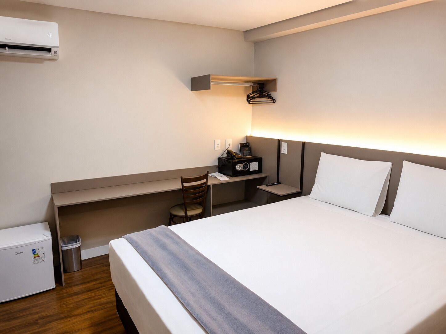 A hotel room with a bed, a desk, a chair, a small trash bin, and an air conditioning unit.