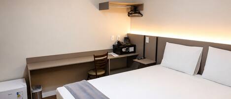 A hotel room with a bed, a desk, a chair, a small trash bin, and an air conditioning unit.