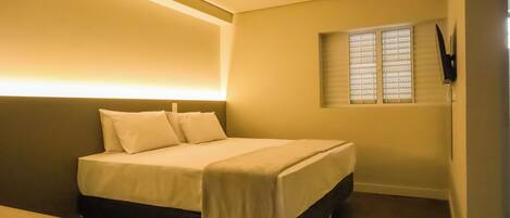 A neatly made bed with white linens in a hotel room with a modern design, including a flat-screen TV mounted on the wall and a window with blinds.