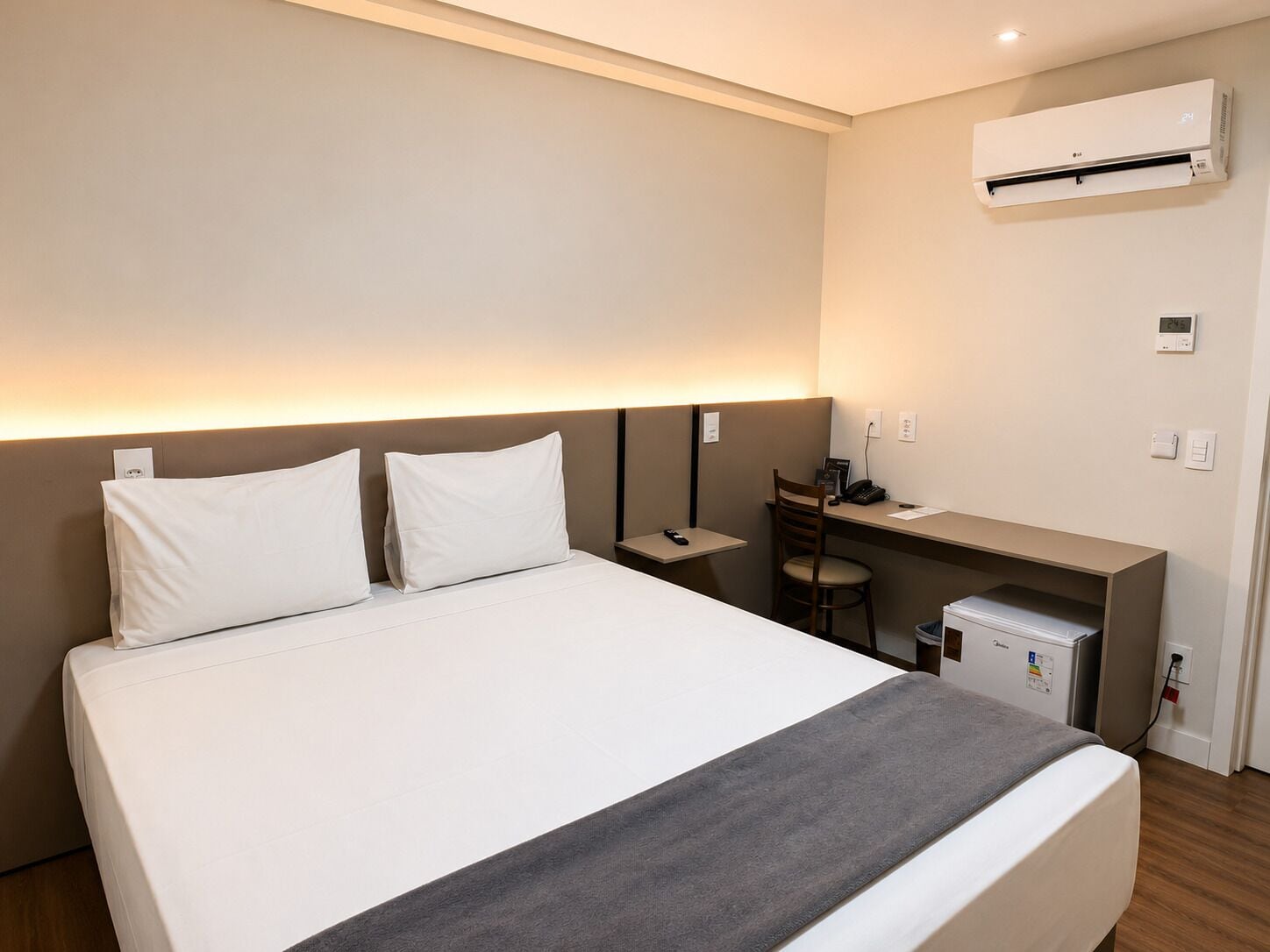 Quarto de hotel com cama, escrivaninha, cadeira, ar condicionado e um pequeno frigorífico.