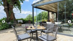 Eine Terrasse im Freien mit einem wintergartenartigen Essbereich, Korbmöbeln und einem großen Baum.