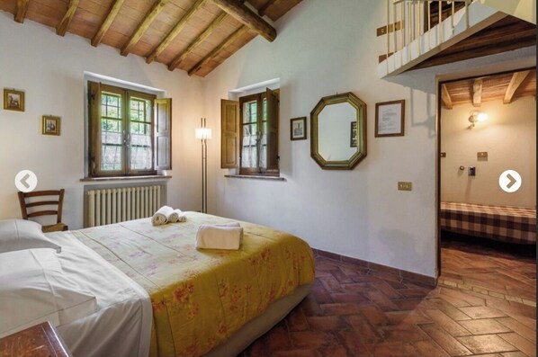 Une chambre à coucher comprenant un lit, deux fenêtres, un radiateur, un miroir et un escalier menant à un étage supérieur.