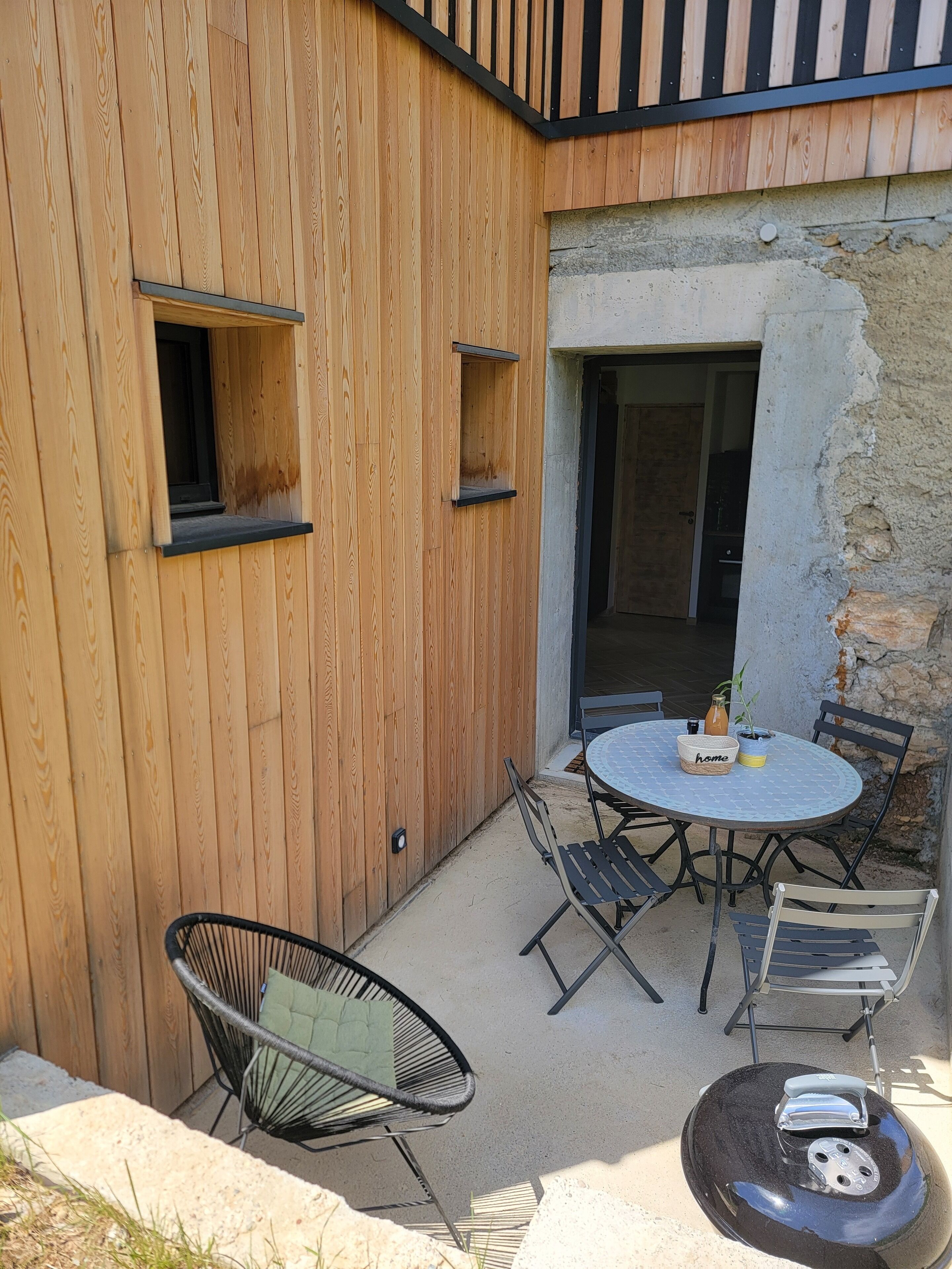En byggnad med träfasad och en liten uteplats med ett runt bord, stolar och en grill.