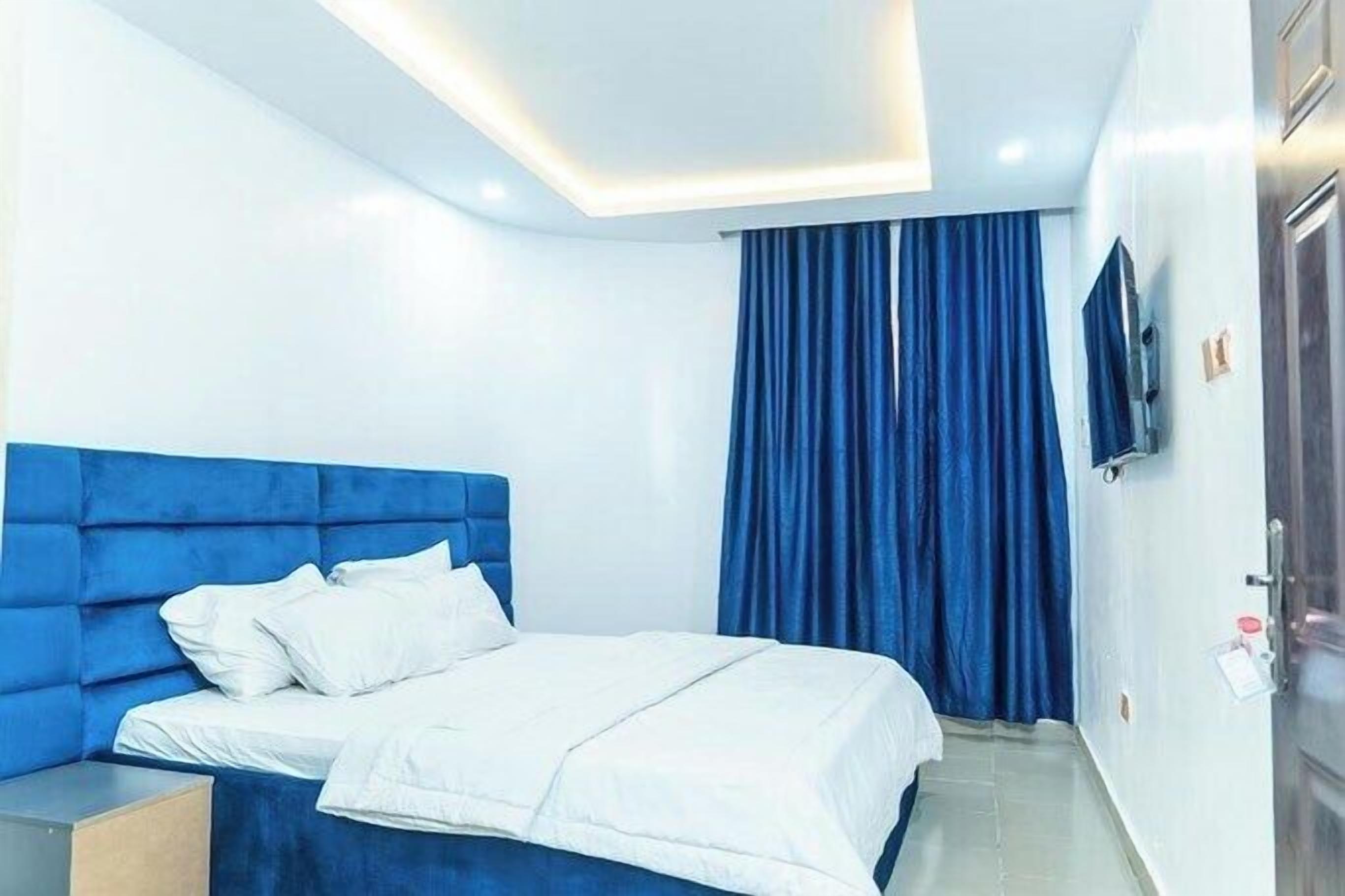 A modern hotel room with a blue headboard, white bedding, and a large window with blue curtains.