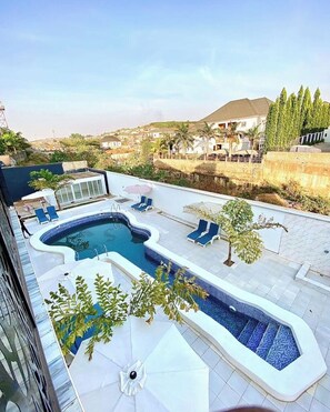 A rooftop pool with a unique shape, surrounded by lounge chairs and umbrellas, set against a backdrop of residential buildings and greenery. - Smart Emmanuel's Place (Abuja)