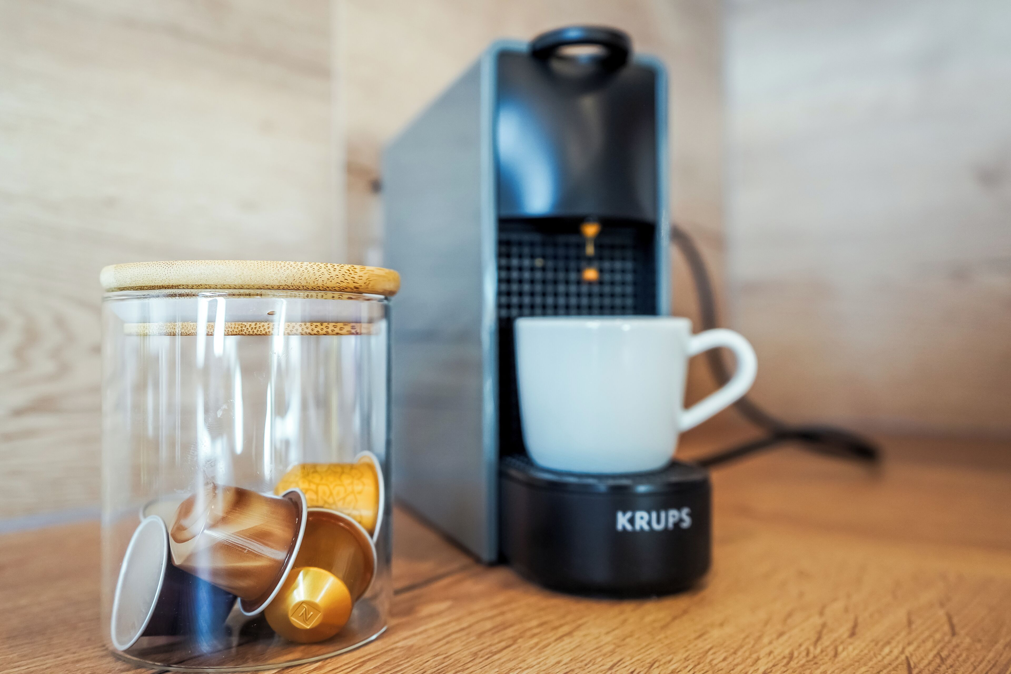 A coffee maker, a glass jar with coffee cups, and a white cup on a wooden surface.