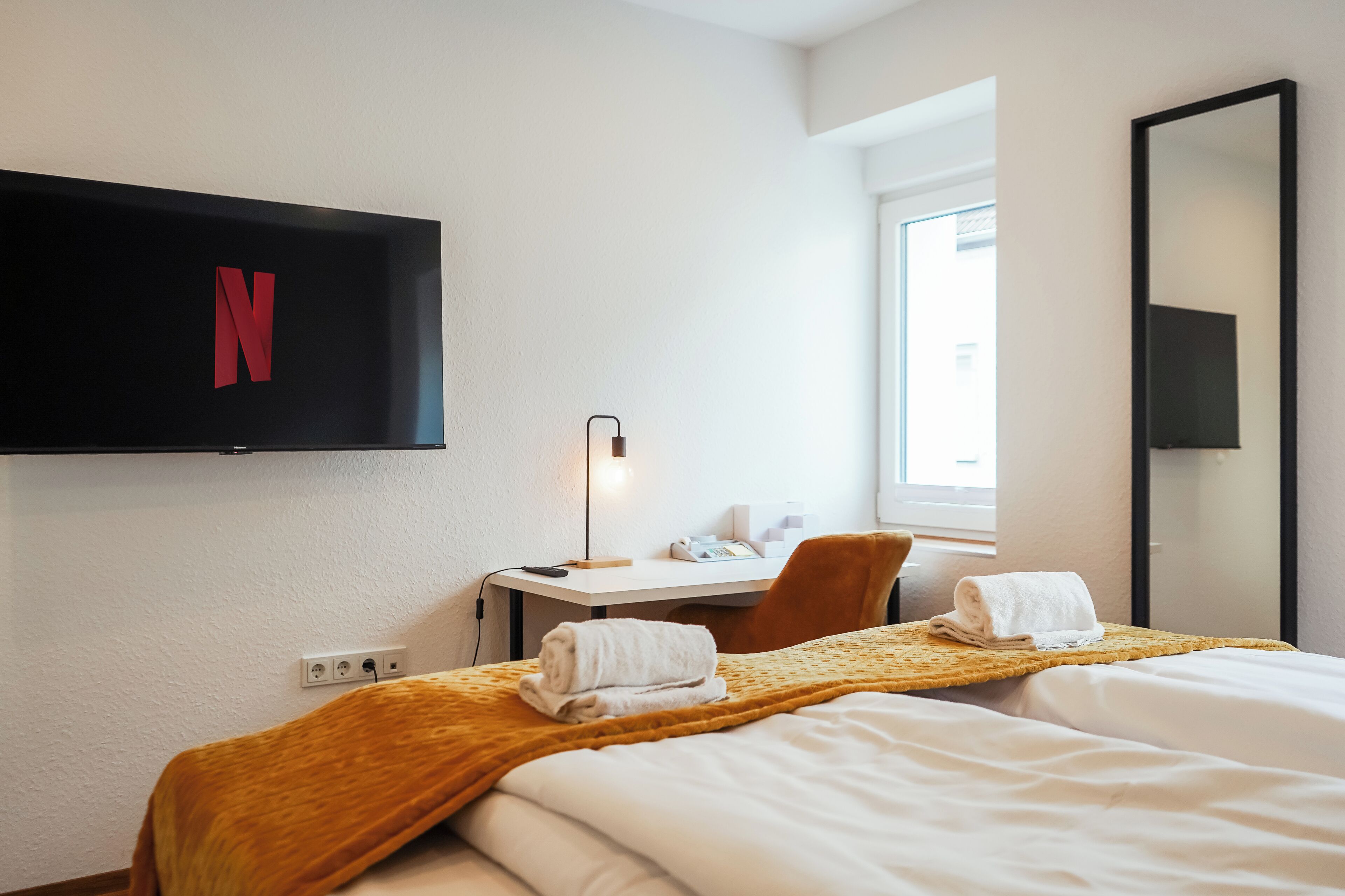Ett hotellrum med en säng, ett skrivbord, en stol, en tv som är monterad på väggen och ett fönster.