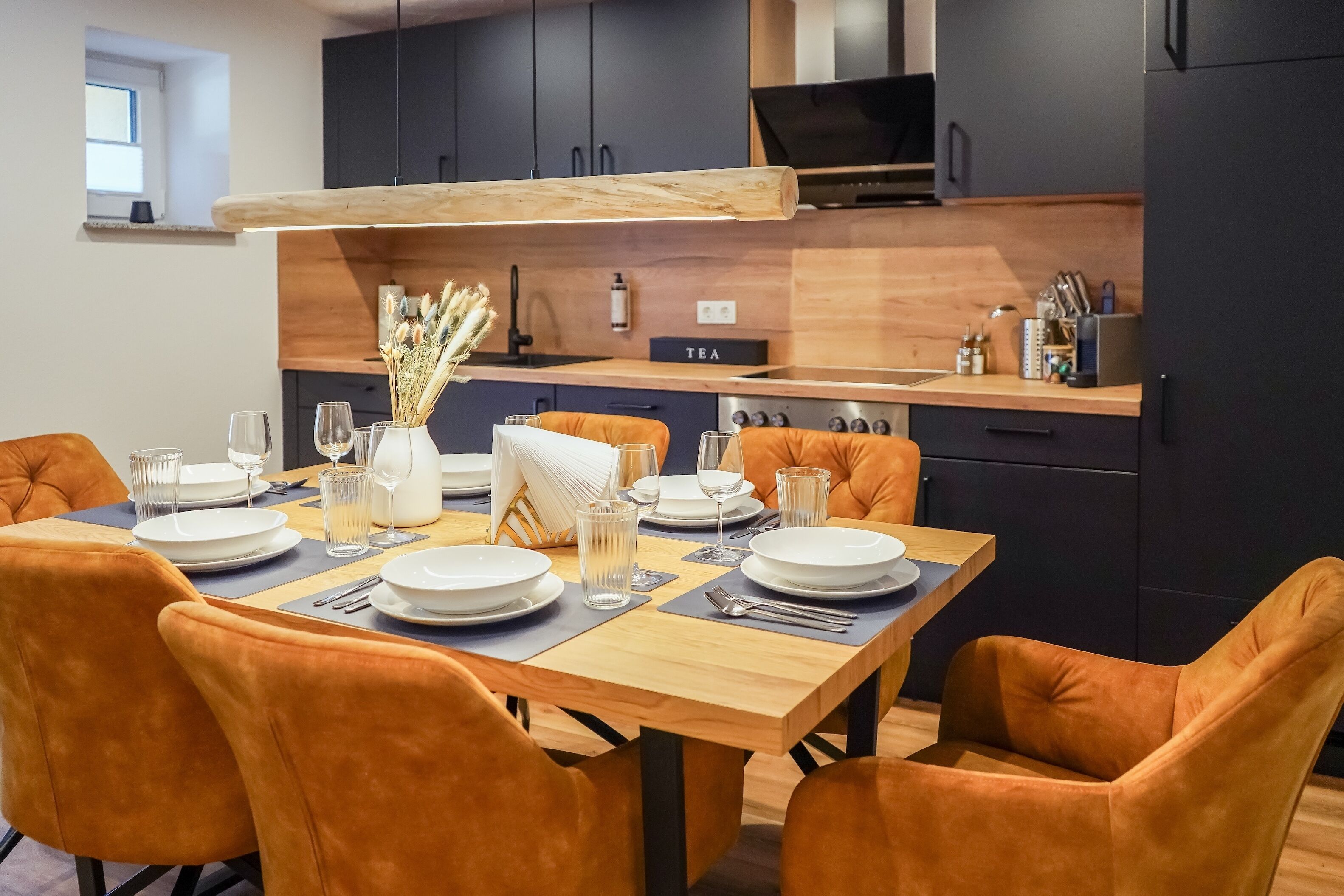 Eine moderne Küche mit dunklen Schränken, einer Holz Arbeitsplatte und einem Essbereich mit orangefarbenen Stühlen und einem Holztisch für vier Personen.
