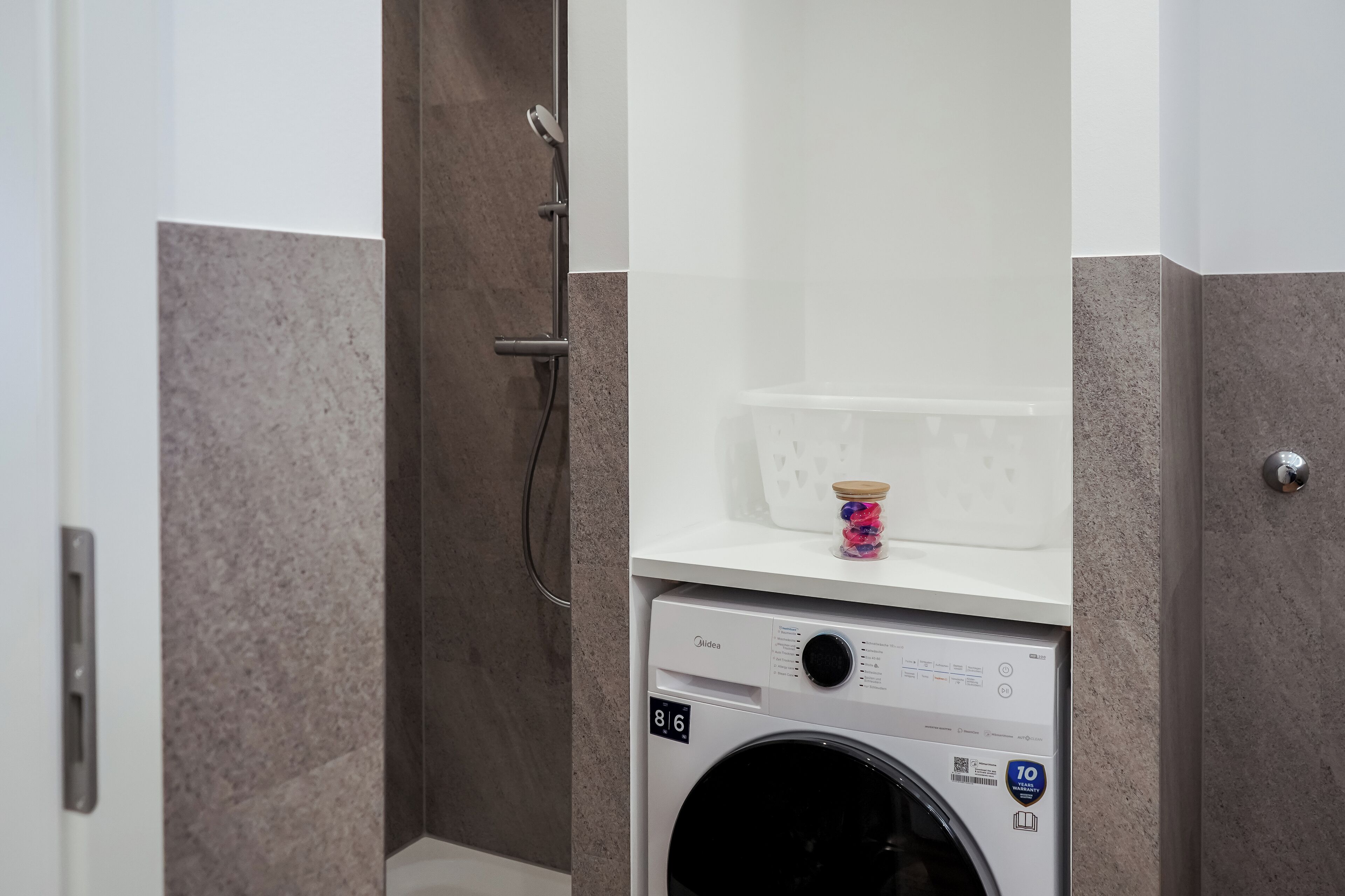 Ein kleines Badezimmer mit Waschmaschine, Dusche und einer kleinen Ablage mit einem Behälter.
