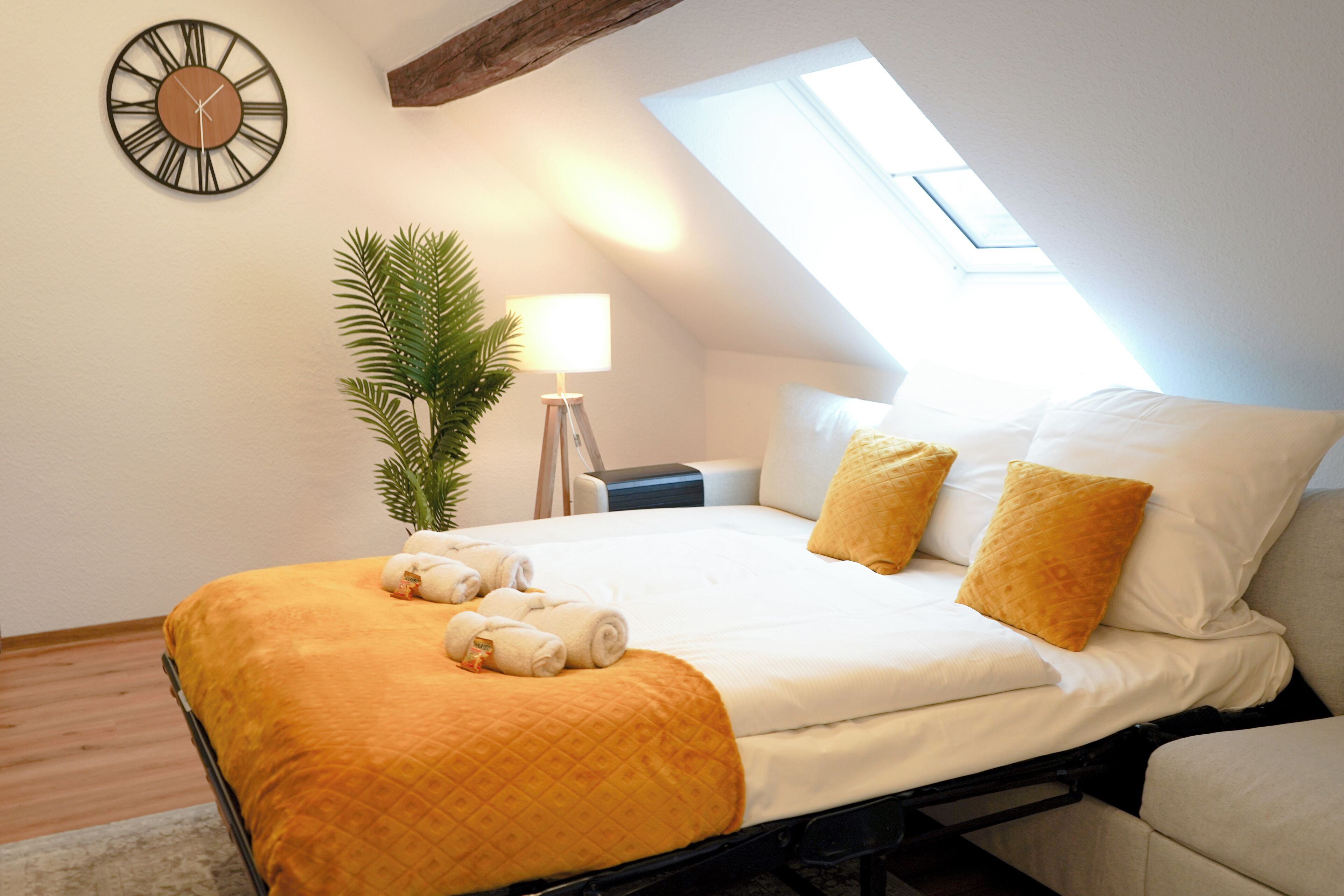 A neatly made bed with a yellow blanket, two pillows, and rolled towels. There is a clock on the wall and a lamp beside the bed.