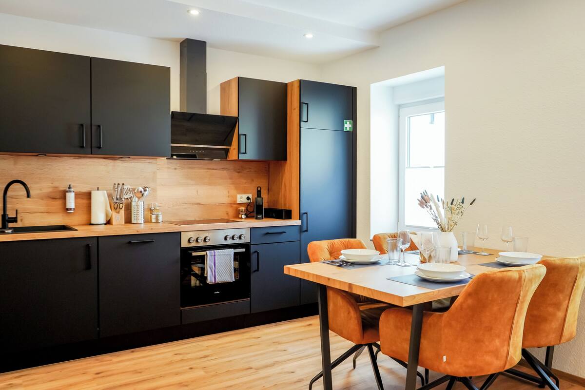 A modern kitchen with dark cabinets, a wooden countertop, and a dining area with orange chairs.