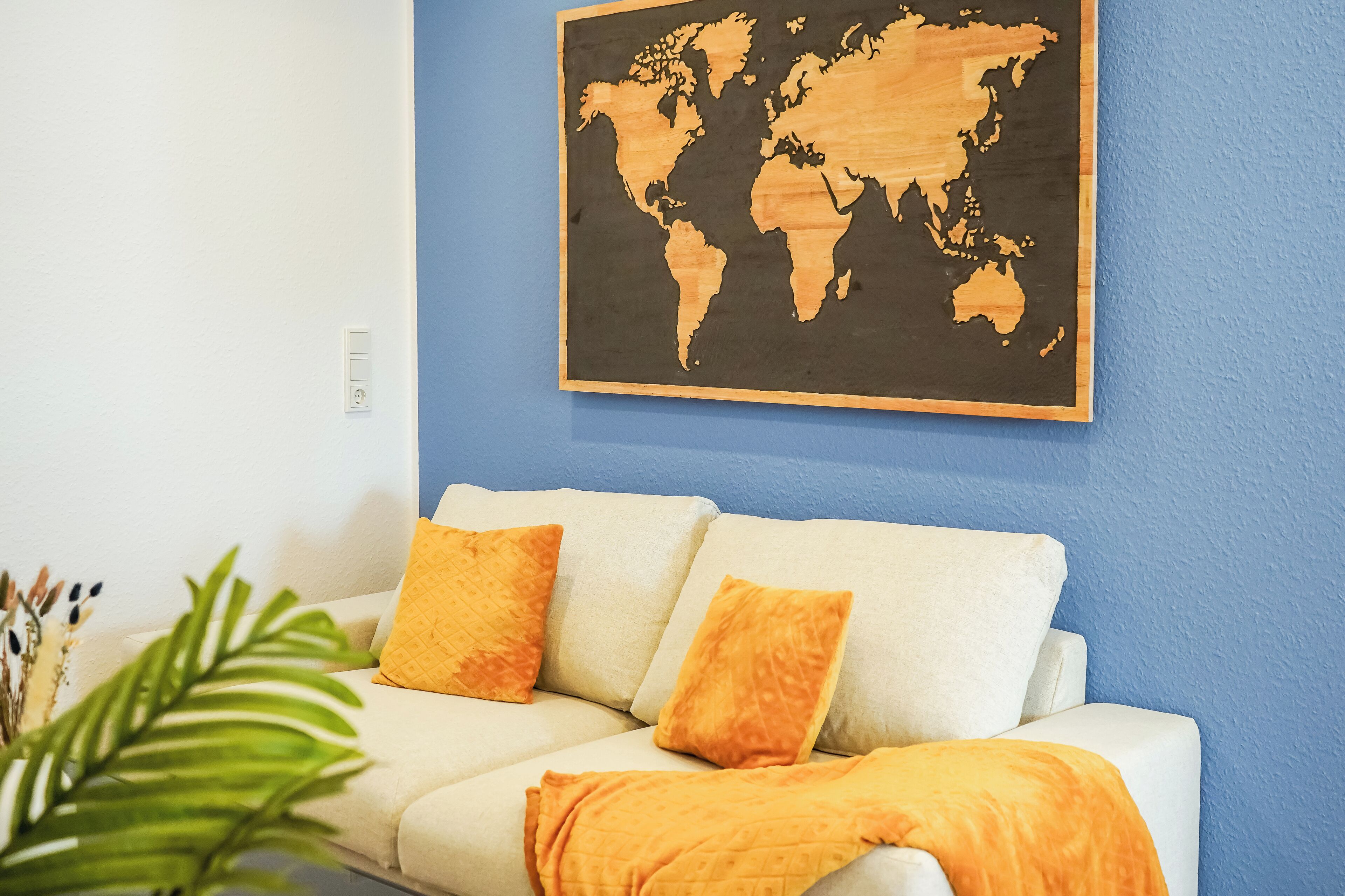A white sofa with two orange cushions and a blanket, against a blue wall with a wooden world map.
