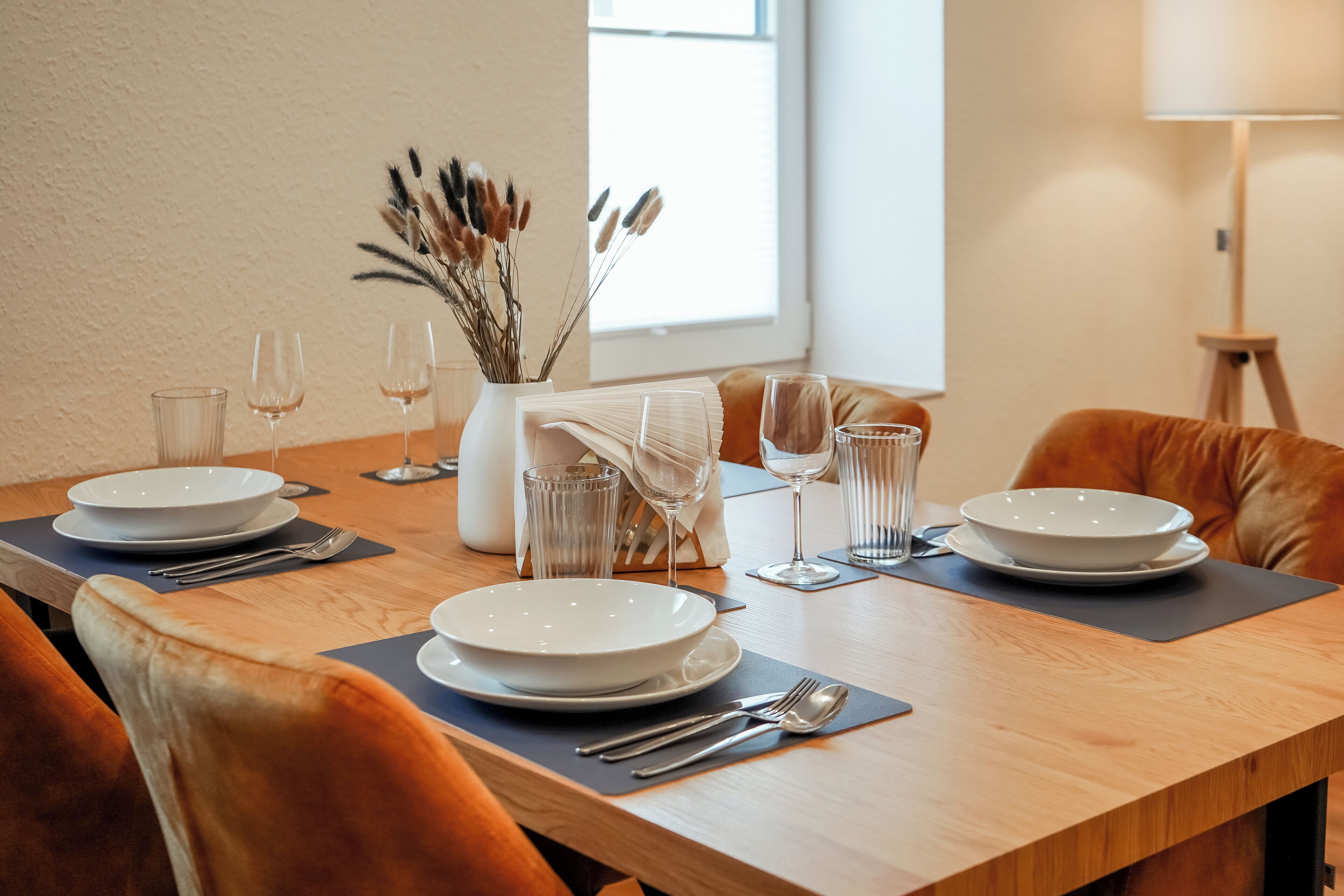 A dining table set with white plates, cutlery, and glasses, accompanied by a vase of dried flowers.