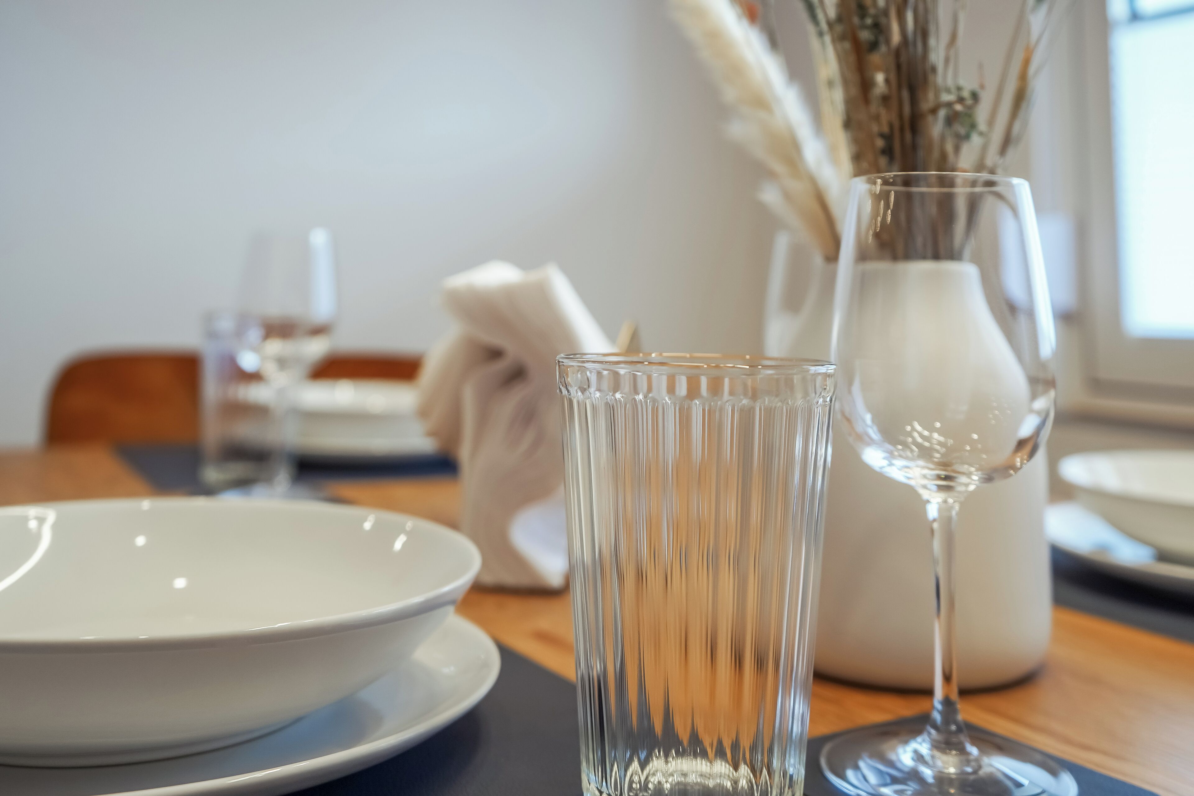 A dining table with a white plate, a clear glass, and a vase with dried plants.