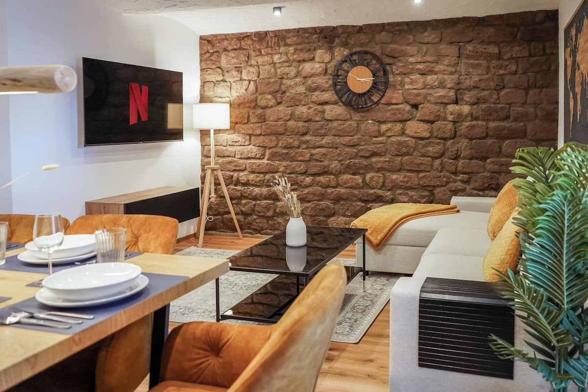 A modern living room with a stone wall, a mounted TV, a clock, and a dining area with a table set for two.
