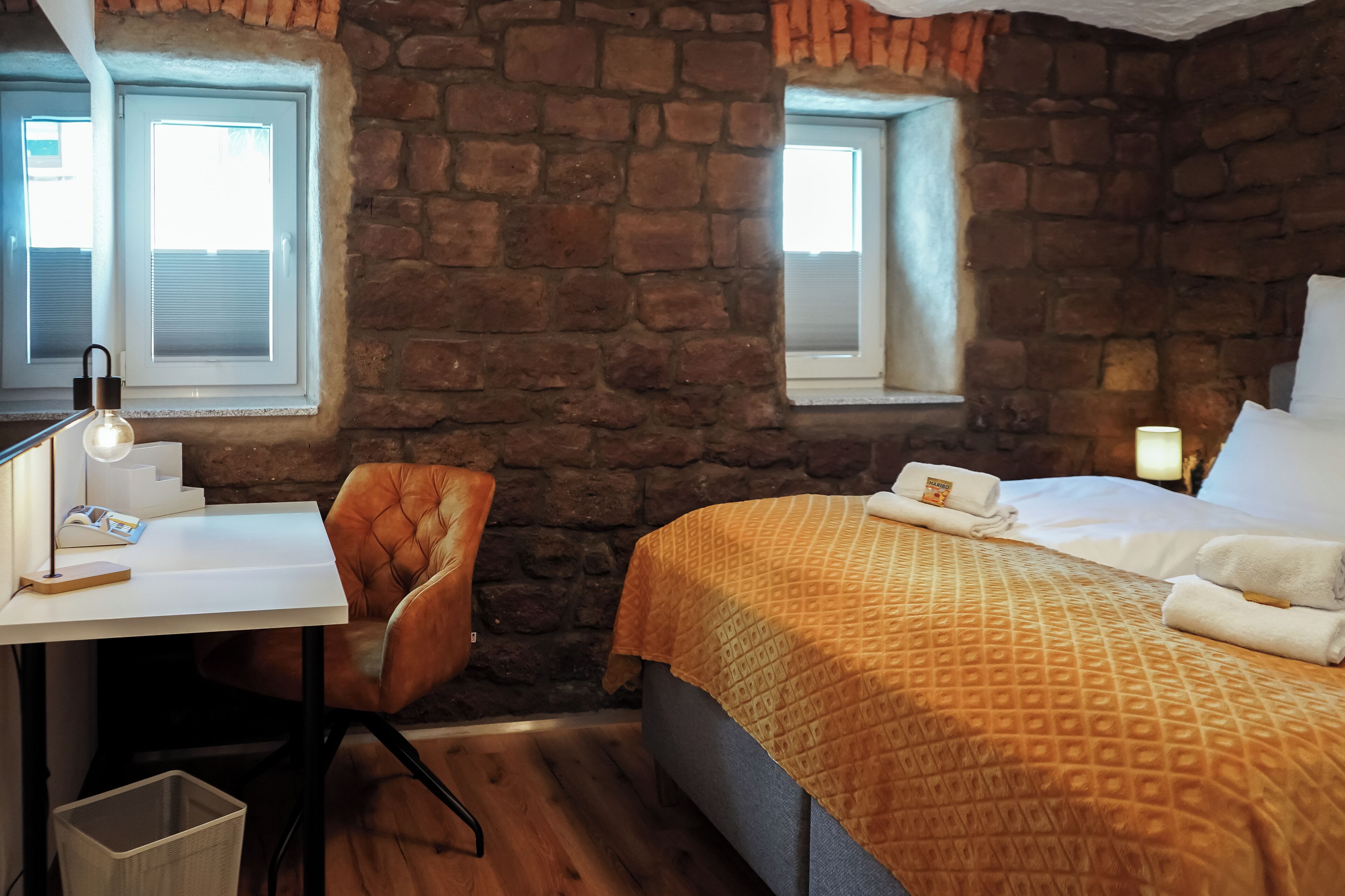 A room with a stone wall, a bed with a patterned blanket, a desk with a lamp, and a chair.