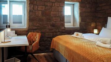 A room with a stone wall, a bed with a patterned blanket, a desk with a lamp, and a chair.