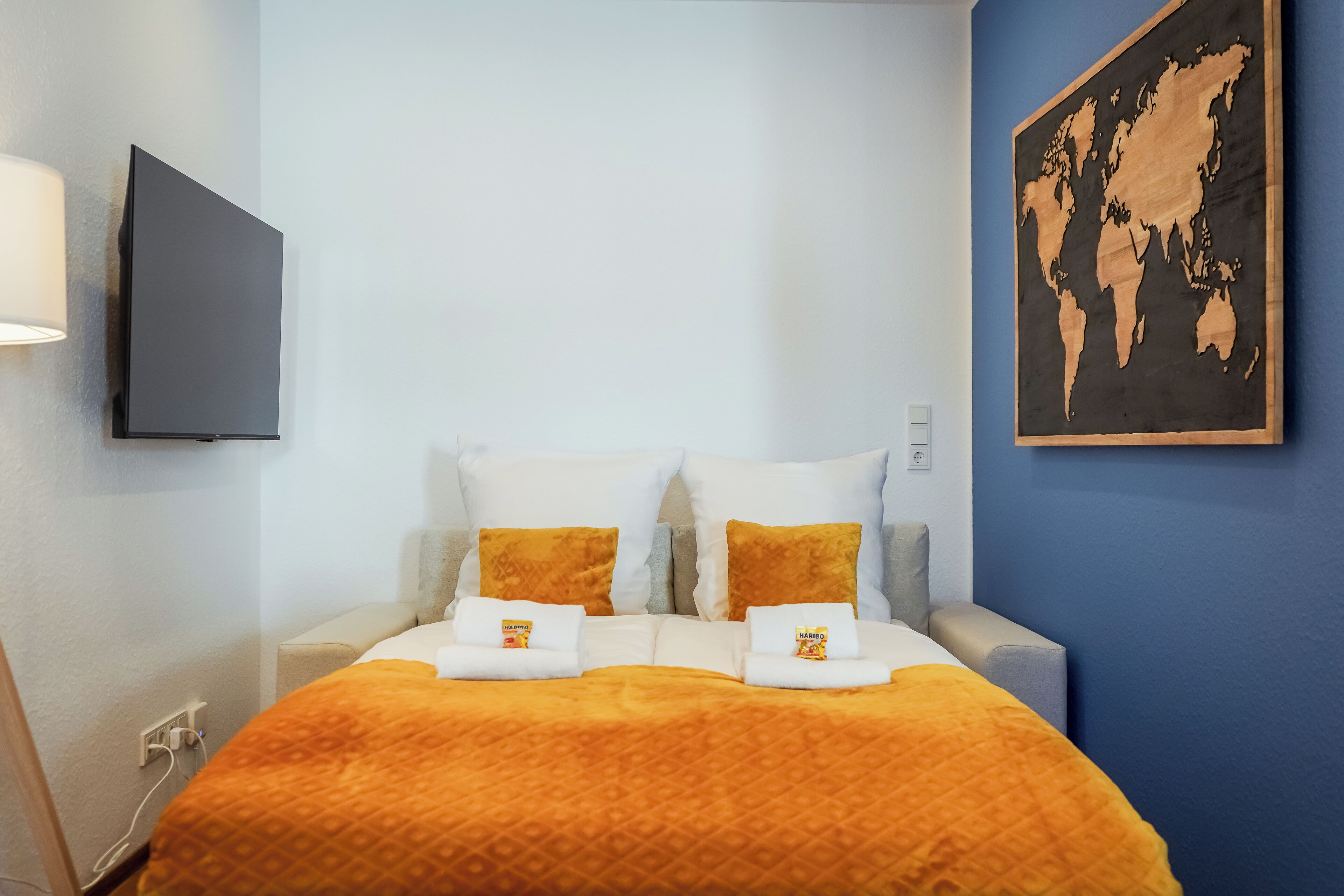 A hotel room with a bed, two pillows, a blanket, a world map on the wall, and a TV mounted on the wall.