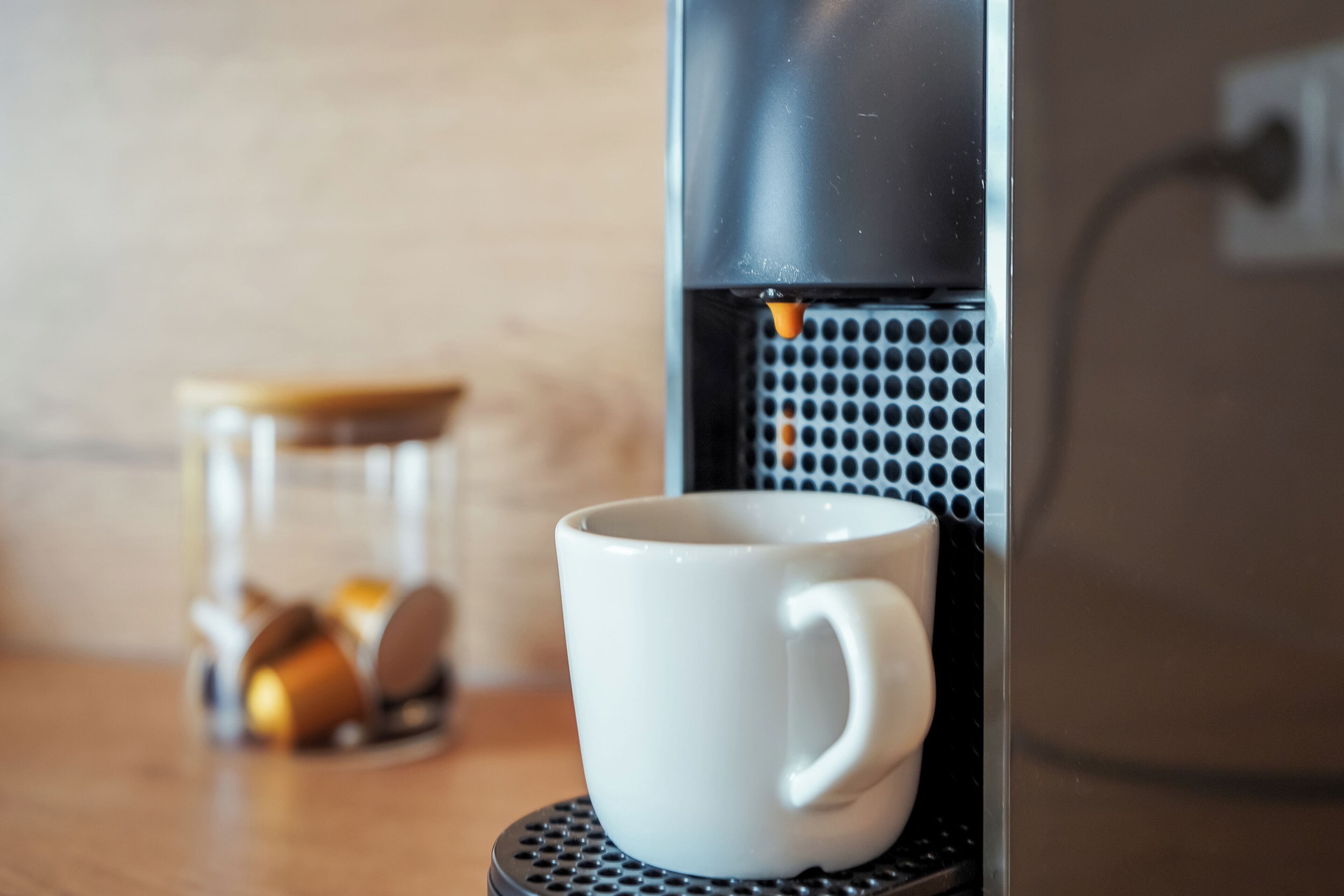 Una cafetera con una taza sobre un posavasos, un tarro de cristal con tapa, y un recipiente metálico con monedas.
