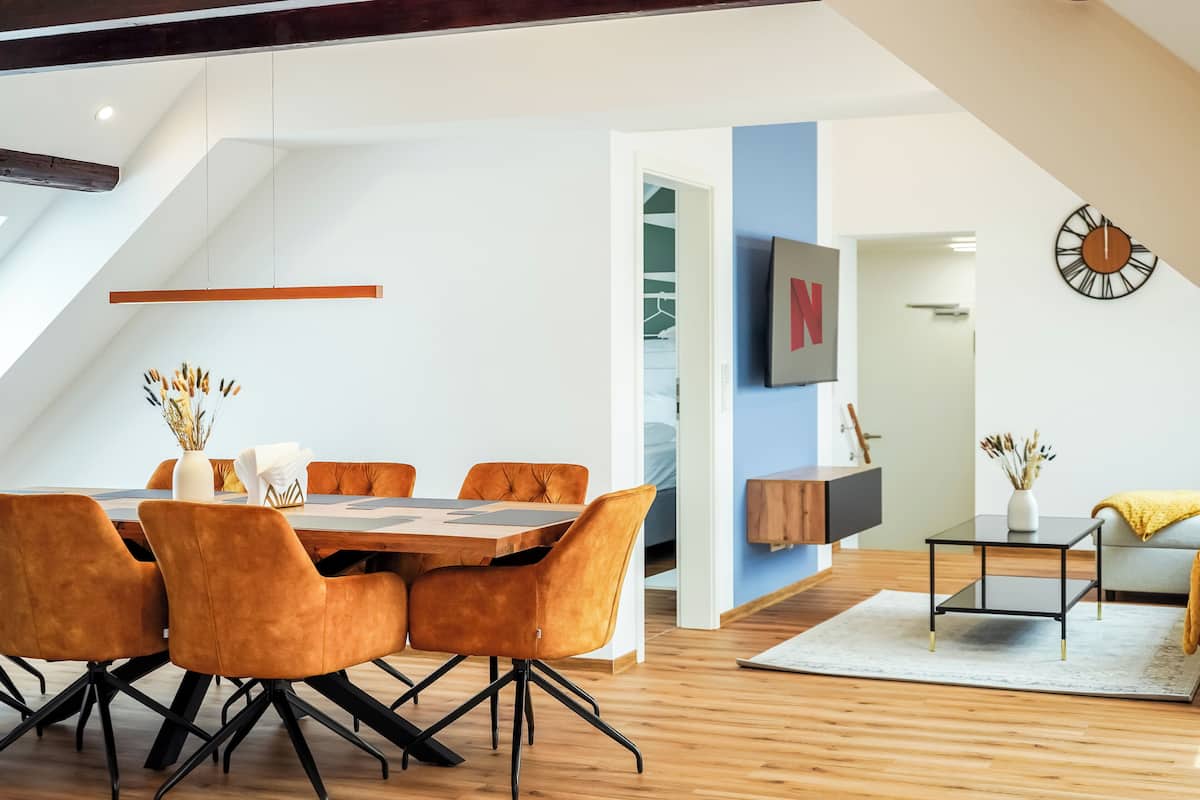 A modern dining area with a wooden table and orange chairs, a TV mounted on the wall, and a clock.