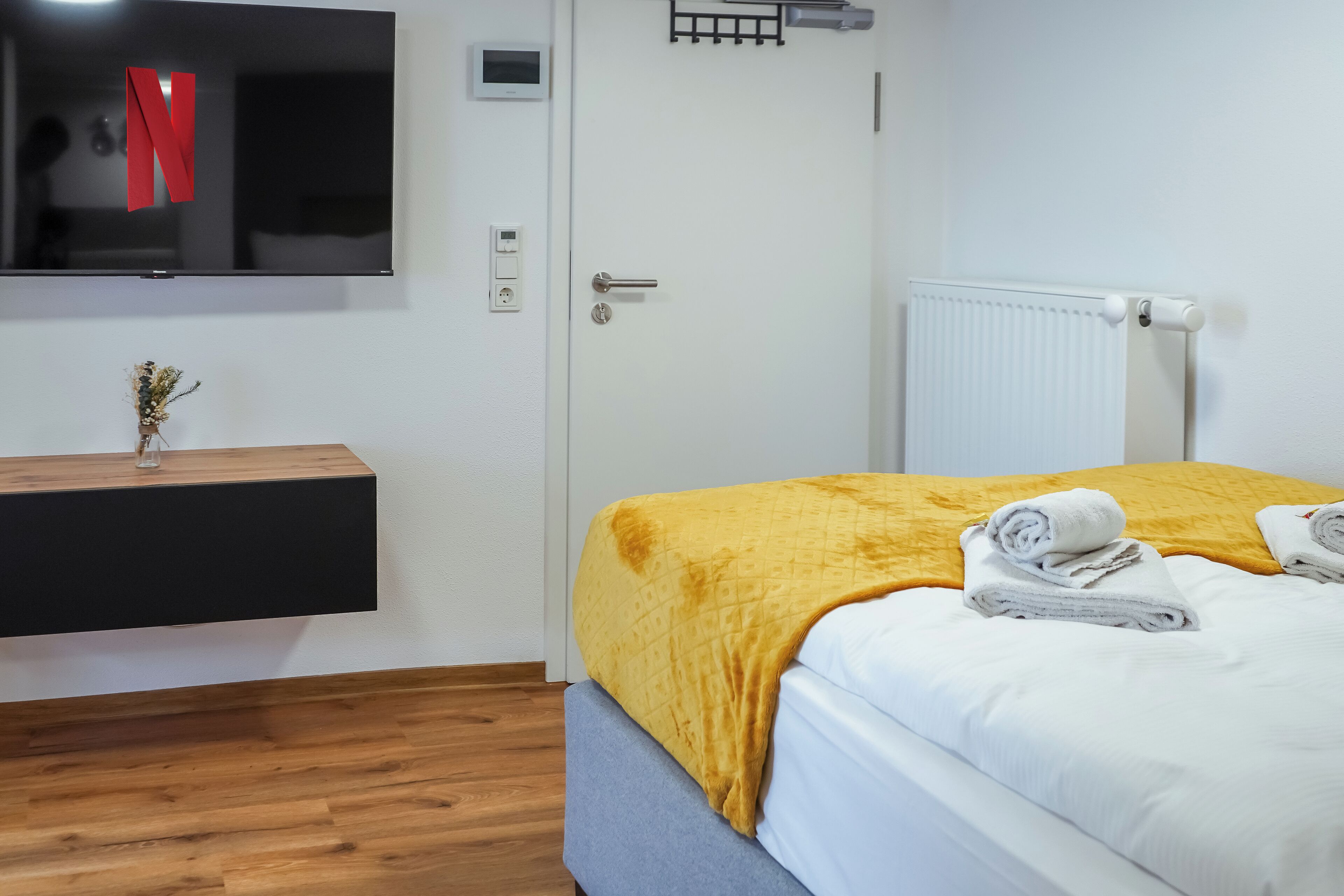 A hotel room with a bed, a TV mounted on the wall, a white radiator, and a wooden floor.