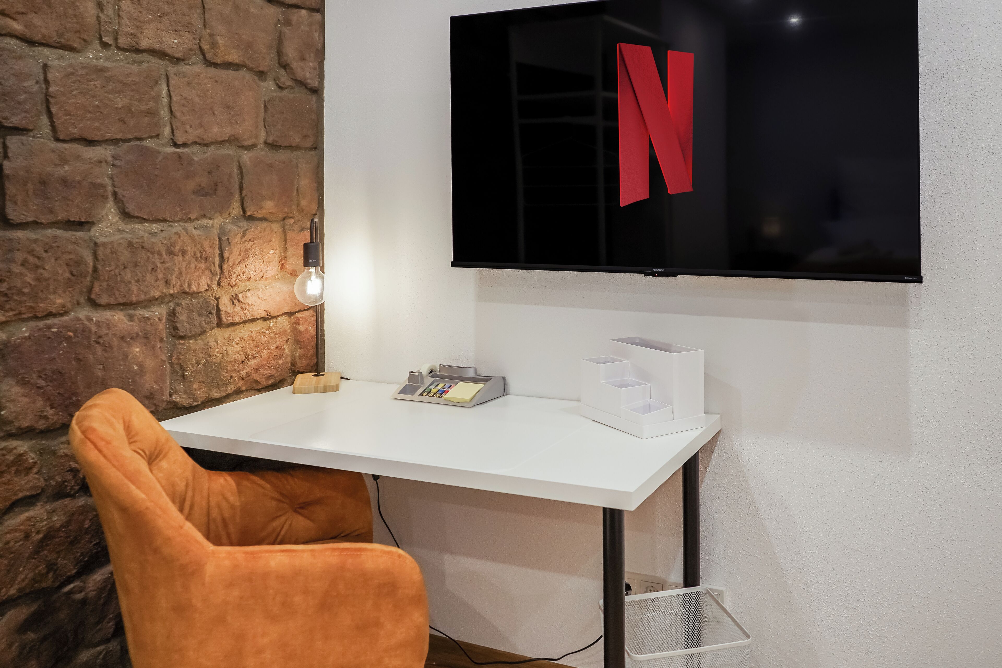 Una habitación con un escritorio blanco, un televisor empotrado en la pared mostrando el logotipo de Netflix, una pared de ladrillo y una sola silla naranja.