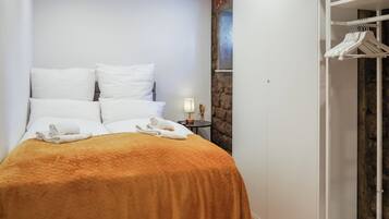 Ein ordentlich bezogenes Bett mit weißen Laken und einer braunen Steude, ein kleiner Nachttisch mit einer Lampe sowie ein weißer Kleiderschrank mit hängender Kleidung.