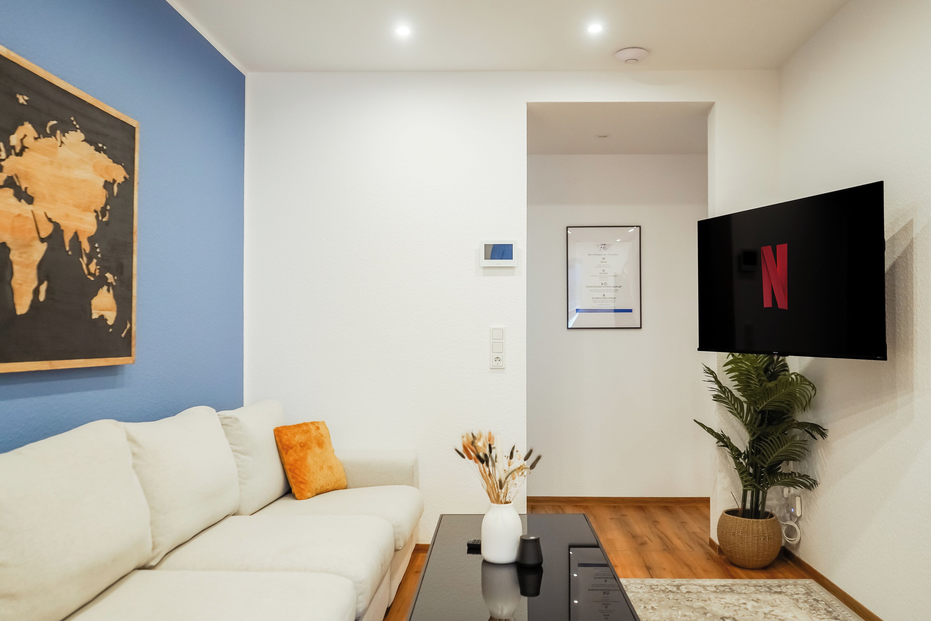 A modern living room with a white sofa, a black coffee table, a world map painting, a plant, and a TV displaying the Netflix logo.
