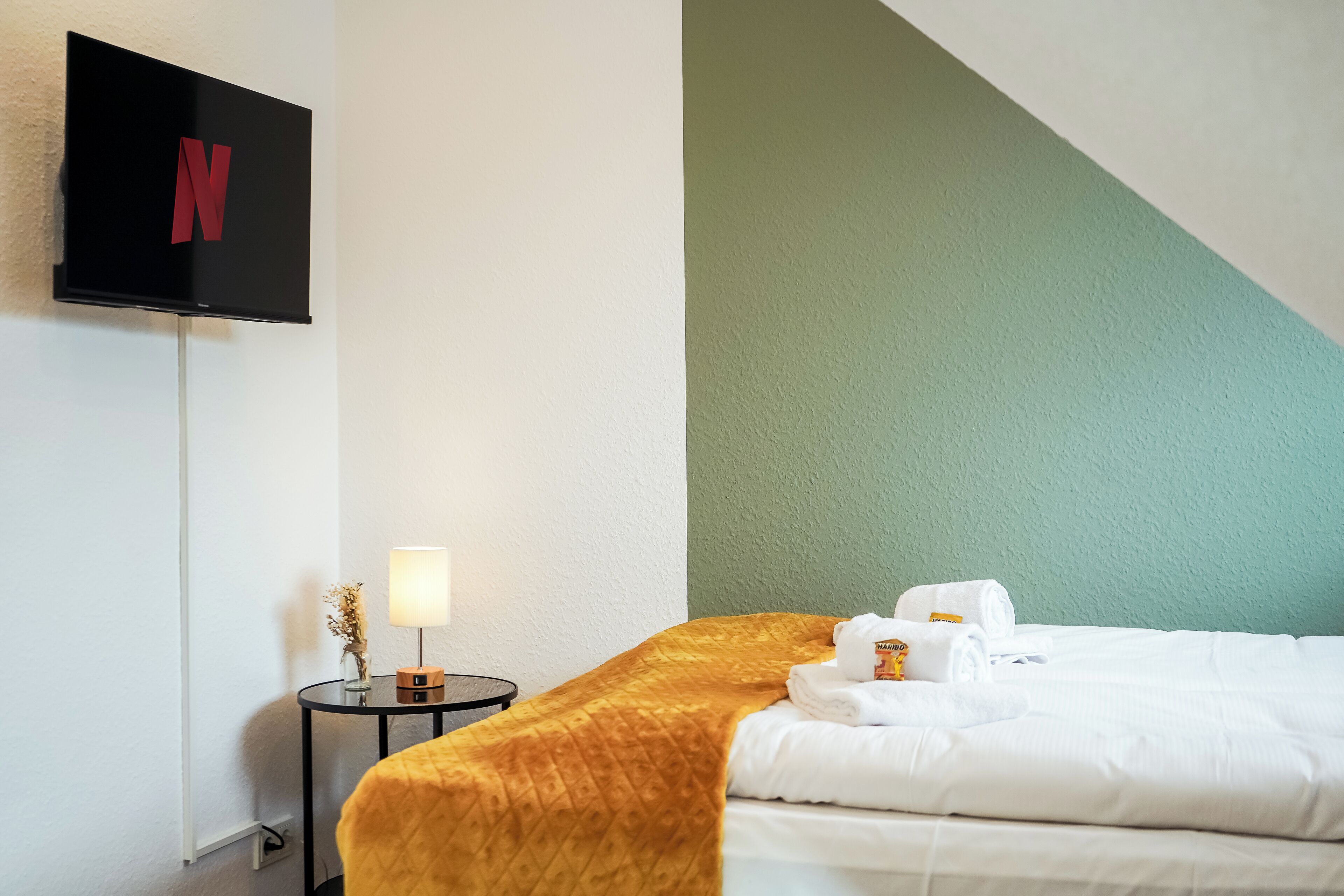 Habitación de hotel con cama, televisor montado en la pared, mesita de noche con lámpara y una pared de acento verde.