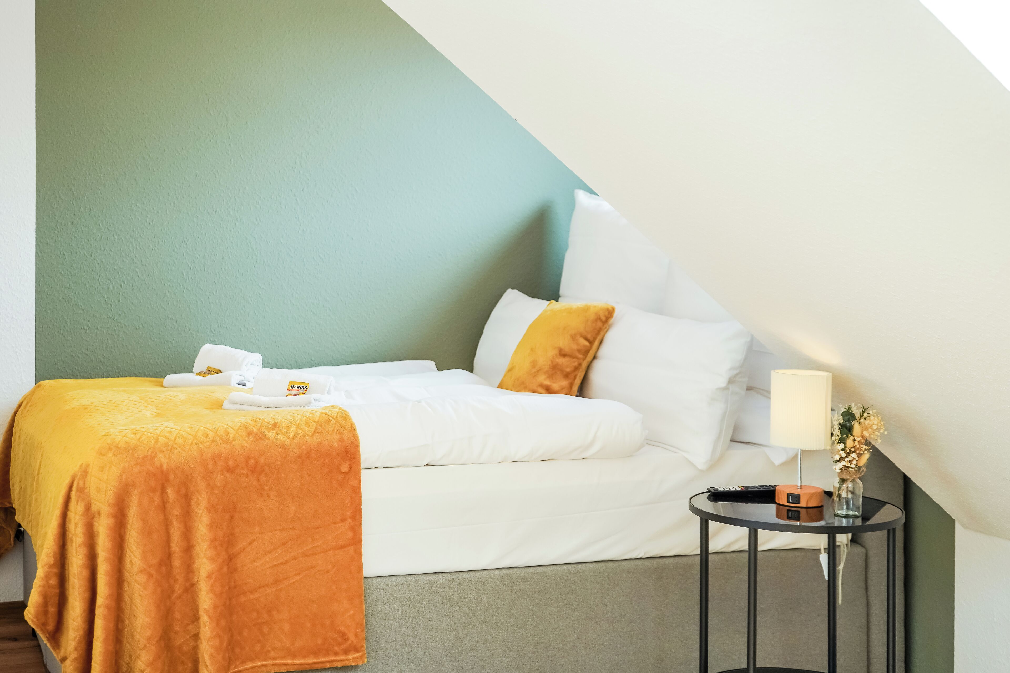 A neatly made bed with a white and orange blanket, a bedside table with a lamp, and a vase with flowers.