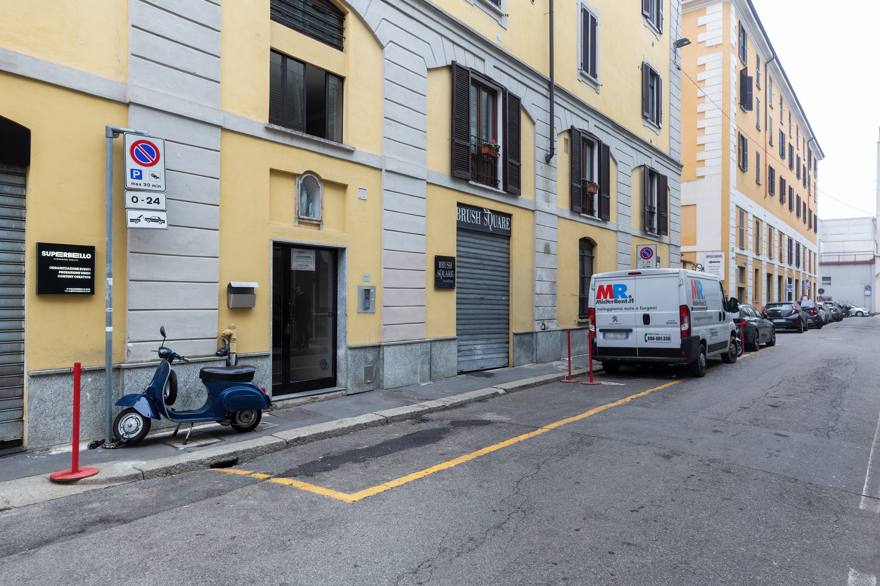 A street with parked cars and a scooter, a no parking sign, and a building with a closed shutter.