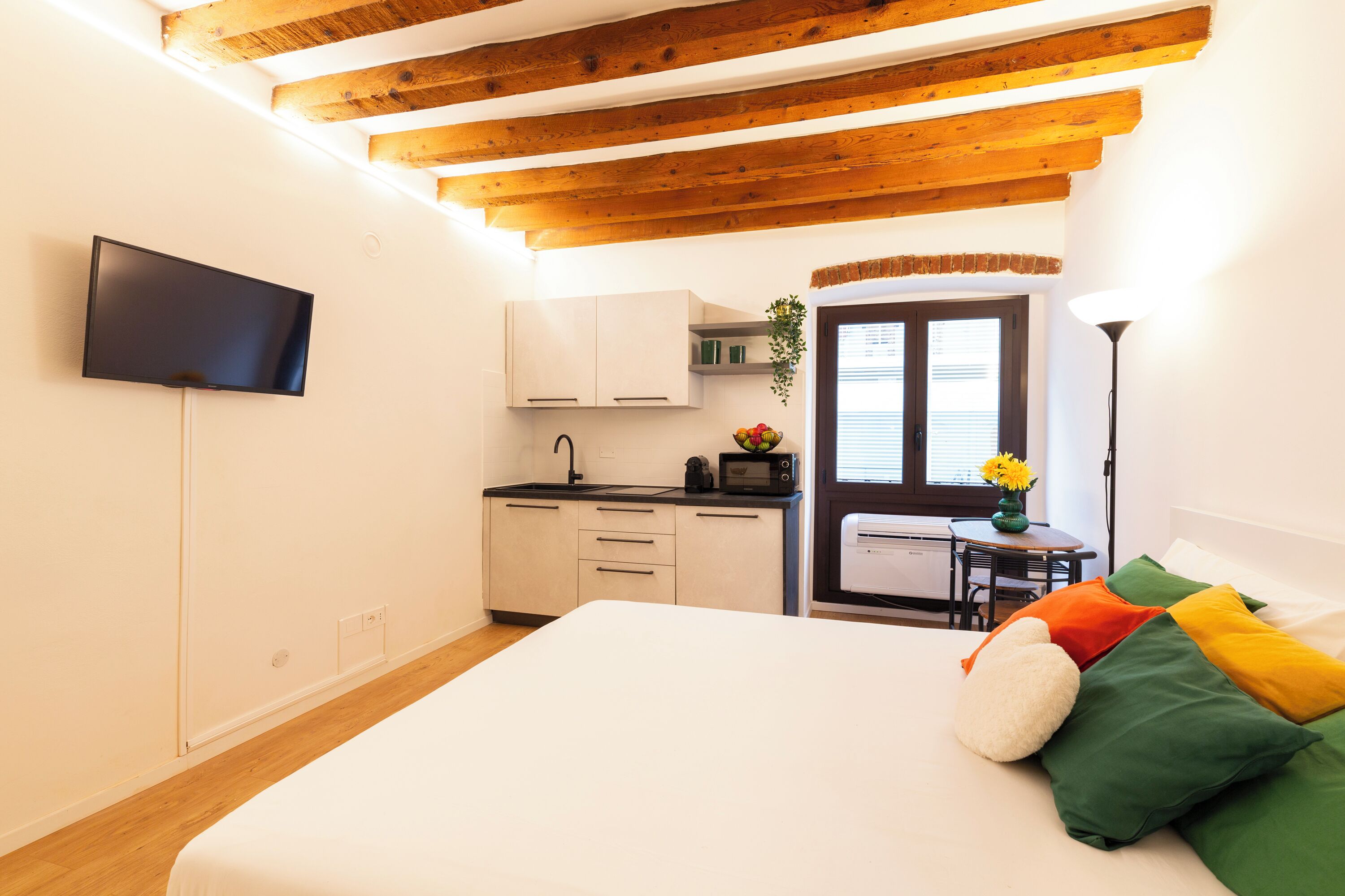 A compact room with a bed, a kitchenette, a TV, and a door leading to another room.