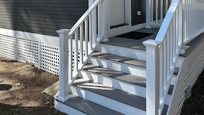 Une petite cabane d’un seul étage, avec une porte d’entrée blanche, une véranda dotée de balustrades blanches et une fenêtre donnant sur les arbres environnants.