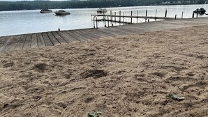 Une plage de sable avec une jetée en bois qui s’avance dans une eau calme. On aperçoit des bateaux au loin.