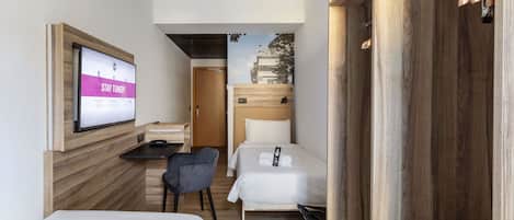 A hotel room with a bed, a desk with a computer, a chair, and a TV mounted on the wall.