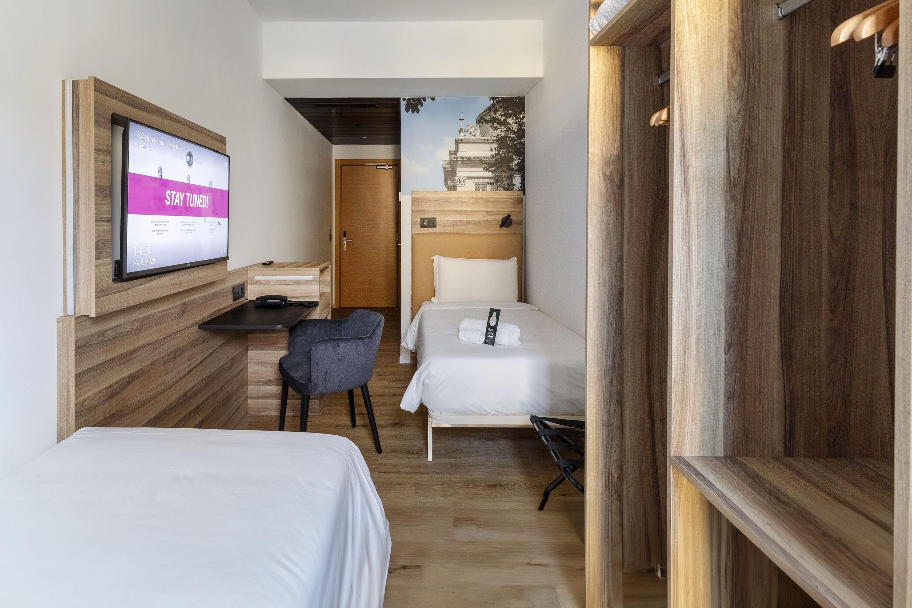A hotel room with two beds, a desk, and a chair. A television is mounted on the wall displaying a message.