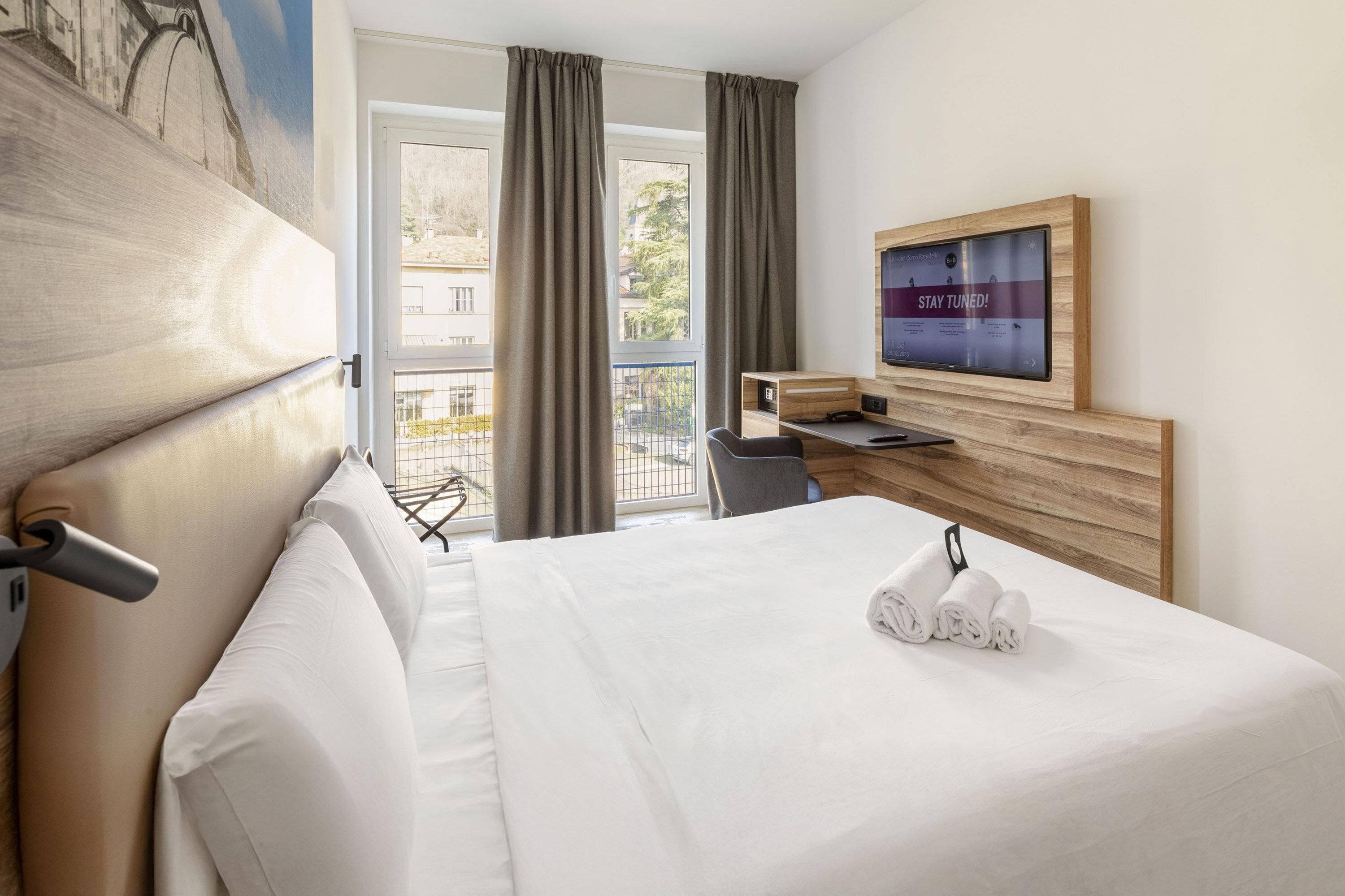 A hotel room with a large bed, a desk, a television, and a balcony with curtains.