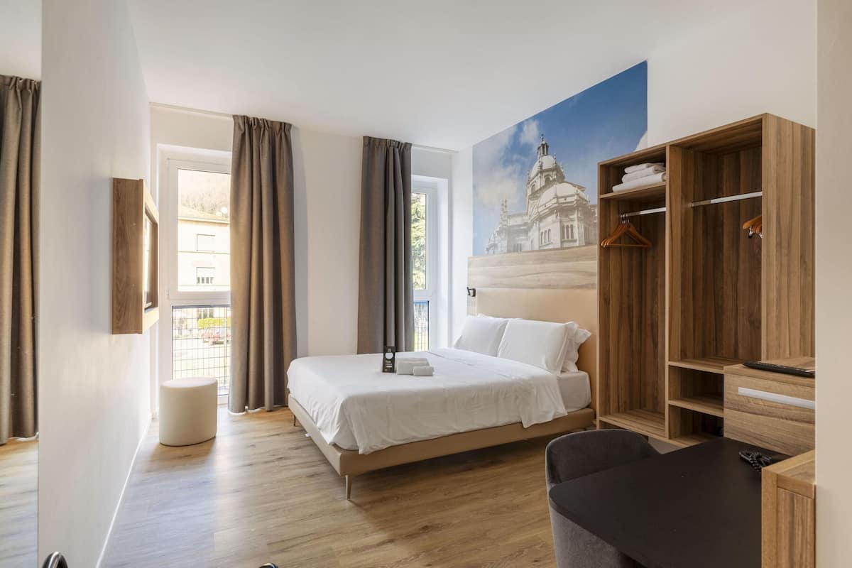 Quarto de hotel moderno com uma cama grande, um guarda-roupa de madeira, uma mesa pequena e vista para um edifício pela janela.