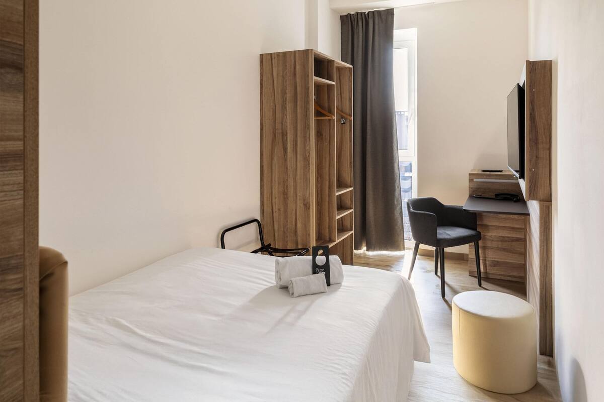 Quarto de hotel com cama, guarda-roupa de madeira, escrivaninha e cadeira, e uma televisão.