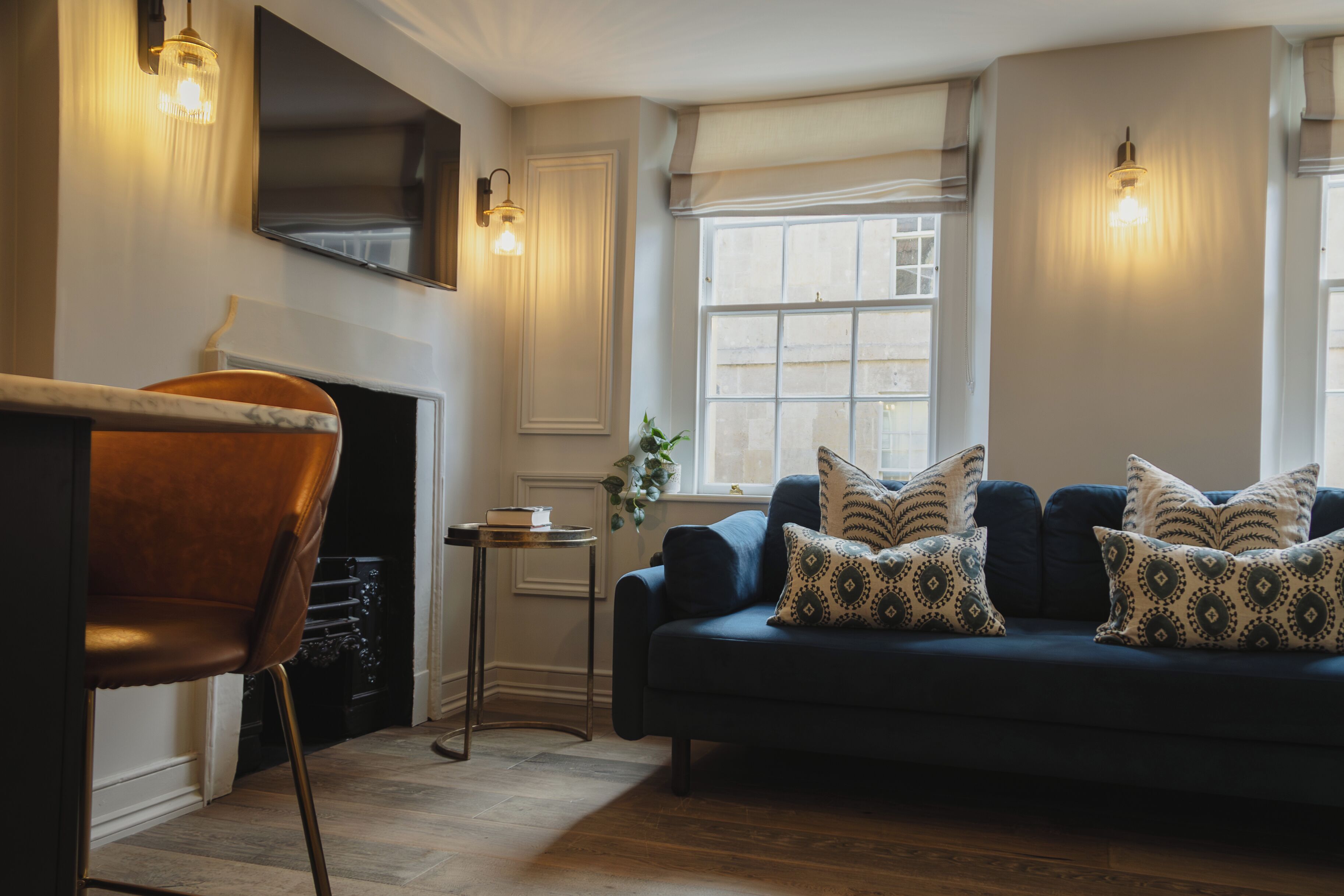 A living room with a dark blue sofa, patterned cushions, a small side table, a wooden chair, a fireplace, and a TV mounted on the wall.