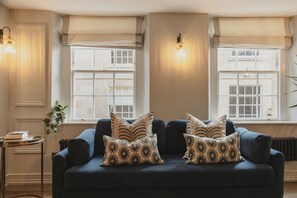 A cozy living room with a dark blue sofa, patterned throw pillows, and two windows with light fixtures. - Princes Street Townhouse (Bath)