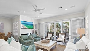 A spacious living room with a large TV, comfortable seating, and a view of palm trees through the windows. - Across from Community Pool and Hot Tub, Close to Beach, Gated Community (Destin)