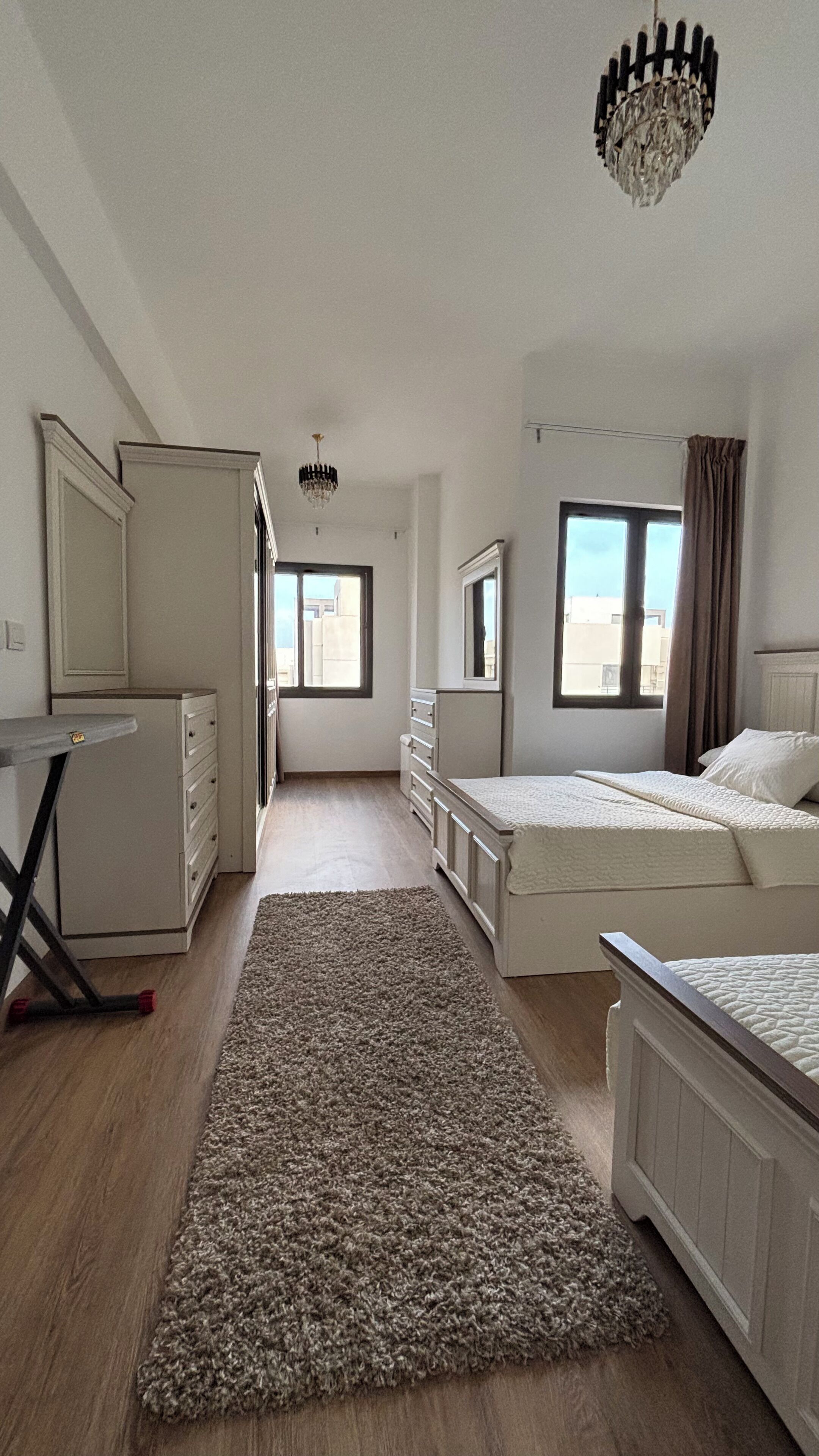 A bedroom with two beds, a desk, a wardrobe, and a view of the outside.