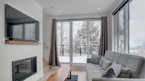 Una sala de estar moderna con un sofá gris, una mesa de centro de madera, una chimenea y ventanas amplias que ofrecen vistas a árboles cubiertos de nieve.